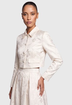 Frau mit nach hinten gegelten Haaren, die eine cremefarbene, floral bestickte, kurze Jacke und einen plissierten Rock vor einfachem Hintergrund trägt.