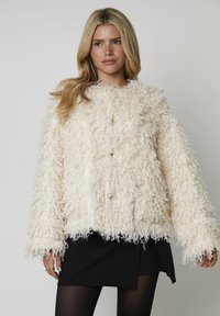 Kremowa, puszysta, krótkia kurtka z teksturowanym wzorem, z dużymi guzikami i szerokimi rękawami, nałożona na czarną spódnicę.