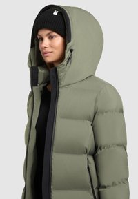 Olijfgroene donzen jas met hoge kraag, ritssluiting en zwarte bies. Voorzien van een grote capuchon en doorgestikte structuur voor isolatie.