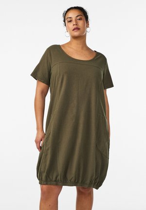 Kvinde iført en løs olivengrøn kortærmet kjole med rund udskæring og elastisk kant, stående mod en hvid baggrund.
