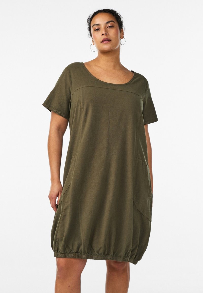 Donna che indossa un vestito corto verde oliva ampio con scollo rotondo e orlo elastico, in piedi davanti a uno sfondo bianco.