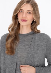 Cardigan grigio con colletto a scialle, maniche lunghe, morbido al tatto e design aperto. Indossato sopra un top a girocollo abbinato.