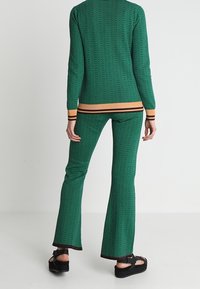 Grünes gestricktes Zweiteiler-Outfit mit Fischgrätenmuster, gerippten orangefarbenen und schwarzen Akzenten, ausgestellten Hosen und flachen Sandalen.
