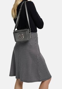 Šedá texturovaná crossbody taška s detailním kulatým logem. Nositelka má na sobě černý top a sukni s chevronovým vzorem, obrácena zády.