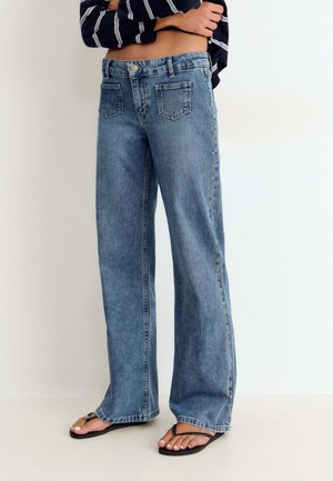 Mujer con vaqueros azules de corte ancho con botones laterales y chanclas negras, de pie frente a un fondo blanco liso.