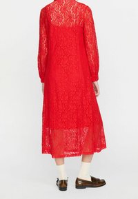 A red lace dress with long sleeves and a scalloped hem, featuring a paisley pattern. The fabric is semi-transparent, layered over a skirt.