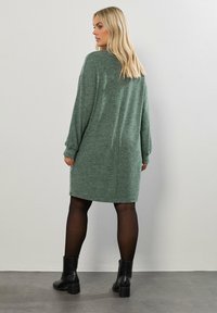 Abito verde lavorato a maglia con maniche lunghe, tessuto testurizzato e vestibilità rilassata, abbinato a stivaletti neri alla caviglia e collant neri trasparenti.