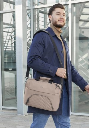 Mann trägt einen blauen Mantel und einen beigen Rollkragenpullover, trägt eine beige Umhängetasche und geht draußen vor einem modernen Gläsernen Gebäude.