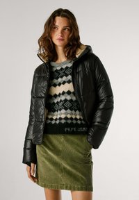 Zwarte pufferjack met een glanzende afwerking, een tan voering en een ritsluiting, gedragen over een gestreepte trui en een groene corduroy rok.