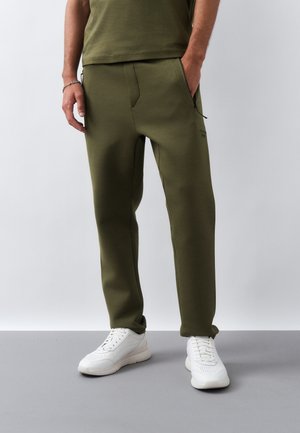 Man die olijfgroene broek en witte sneakers draagt, staat met één hand in zijn zak tegen een effen lichte achtergrond.