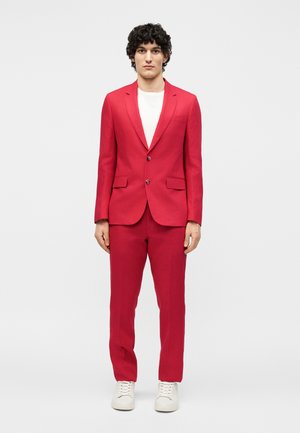 Giovane uomo con capelli ricci in piedi di fronte, indossa un completo rosso, camicia bianca e sneakers bianche su uno sfondo chiaro uniforme.