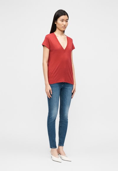 Top vermelho de manga curta com decote em V, corte descontraído, combinado com jeans skinny azuis e sapatos brancos de bico fino. Textura suave e design simples.