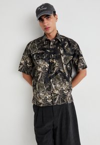 Jeune homme portant une casquette grise et une chemise camo à manches courtes avec un grand motif d'étoile noire, appuyé contre un mur blanc.