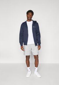 Navy Zip-Hoodie aus Baumwolle, graue Shorts, weißes T-Shirt und weiße Sneakers. Mit Logodetails und einer lässigen Passform.