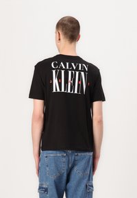 Persona con pelo corto que lleva una camiseta negra de Calvin Klein Jeans y pantalones cargo de mezclilla azules, de espaldas frente a un fondo plano.