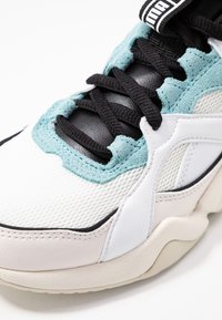 Sneaker bianco realizzato con materiali in rete, suede e pelle. Lacci neri, dettagli verde menta e una suola beige spessa con finiture testurizzate.