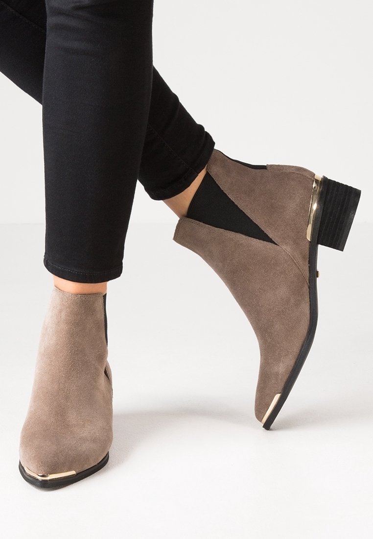 Bottines en daim marron avec côtés élastiques noirs, accents métalliques dorés au talon et à l\'avant, portées avec un pantalon noir ajusté.