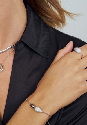 Sølv smykkesæt med en struktureret oval ring og et matchende armbånd med perleaccent, båret på en bar arm imod en sort skjorte.