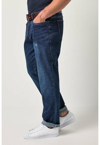 Hombre con vaqueros oscuros rizados, cinturón marrón, zapatillas blancas y una camisa azul marino con una mano en el bolsillo contra un fondo plano.