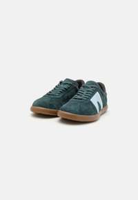 Sneaker in suede verde con un accento bianco sul lato, suola in gomma marrone e lacci piatti. Forma della punta arrotondata e design a profilo basso.