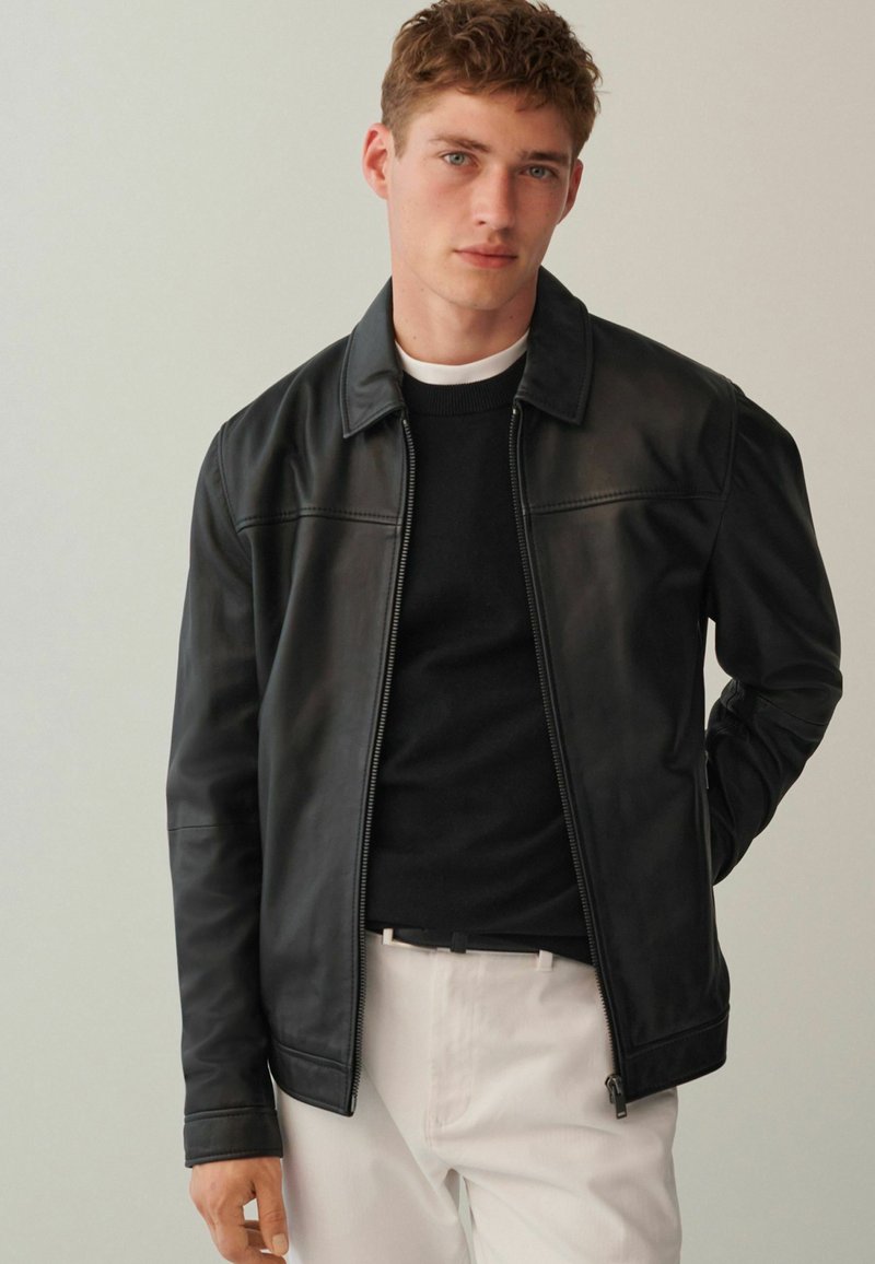 Schwarze Lederjacke mit Reißverschluss, spitzem Kragen und Schulternähten. Trägt man über einem schwarzen Pullover, kombiniert mit hellen Hosen.