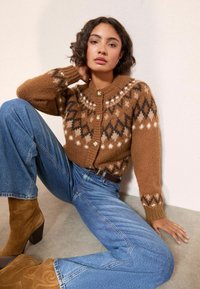 Naine istub vastu seina, seljas mustriga pruun kudum, sinised teksad ja tan värvi nahast kontsakingad, käsi toetatud kaelale.