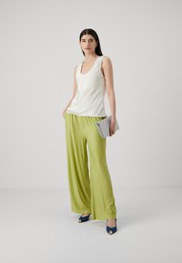 Weißes Satin-Trägershirt mit U-Boot-Ausschnitt, kombiniert mit grünen, plissierten Hosen mit weitem Bein. Silberne Clutch und marineblaue Absätze mit Akzentdetails.