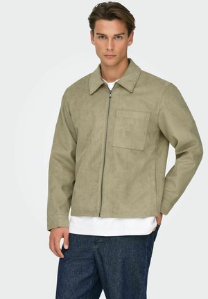 Man draagt een licht olijfgroene jas met rits en borstzak, wit shirt en donkerblauwe spijkerbroek, staande tegen een effen lichte achtergrond.