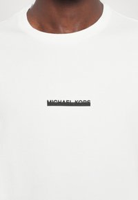 Balta kokvilnas T-kreklā ir melns gumijots "Michael Kors" logos, kas atrodas vidū uz krūtīm. Apaļš izgriezums un īsas piedurknes.