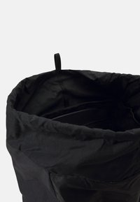 Crna platnena torba s širokim, otvorenim vrhom i petljom za vješanje. Materijal djeluje glatko s nekakvom teksturom, dizajniran za lak pristup.