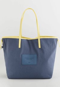 Bolsa de lona marina con ribetes y asas amarillas. Presenta un parche rectangular azul en la parte frontal con texto en relieve. Cierre de cremallera.