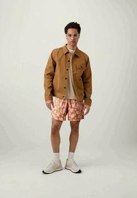 Brown denim jacket, beige t-shirt, pink floral shorts, and white sneakers with gray soles; clear frame glasses. Casual outfit with varied textures.