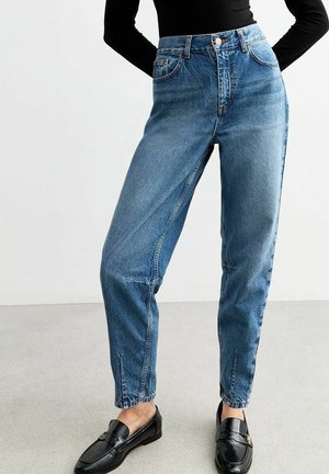 Kvinde iført højtaljede, skrå blå jeans med frontsplits ved anklerne, kombineret med sorte loafers og en sort langærmet top.