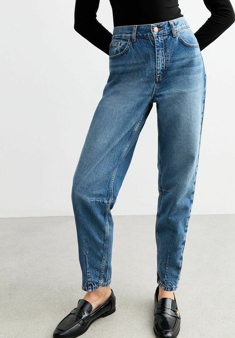Femme portant un jean bleu taille haute, fuselé, avec des fentes à l'avant aux chevilles, associé à des mocassins noirs et un haut noir à manches longues.