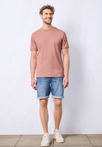T-shirt di cotone rosa chiaro con maniche corte, abbinata a pantaloni shorts in denim blu arrotolati e sneakers beige. Design semplice e casual.
