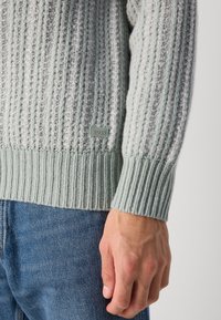Gray knit sweater with ribbed texture and contrasting light gray accents. Features a small logo patch near the hem and ribbed cuffs.