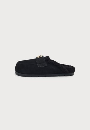 Zapatilla negra tipo slip-on con forro suave y peludo y una sola correa con una hebilla dorada sobre un fondo liso.