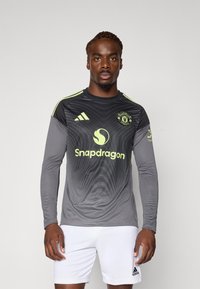 Langärmliges graues Fußballtrikot mit schwarzen geometrischen Mustern, gelben Akzenten und Adidas-Logo. Getragen mit weißen Shorts.