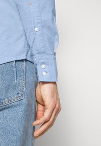 Vaaleansininen napillinen paita, jossa on valkoiset napit ja kevyt tekstuuri, yhdistettynä haalistuneisiin vaaleansinisiin farkkuihin.