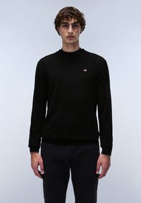 Zwarte gebreide trui met lange mouwen, ronde halslijn en een klein rood-wit vlaglogo op de borst. Draagt een donkere broek.