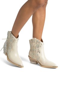 Bottes de cowboy en simili cuir blanc, avec des orteils pointus, des clous décoratifs en argent et des franges. Talon bas empilé et design découpé complexe.