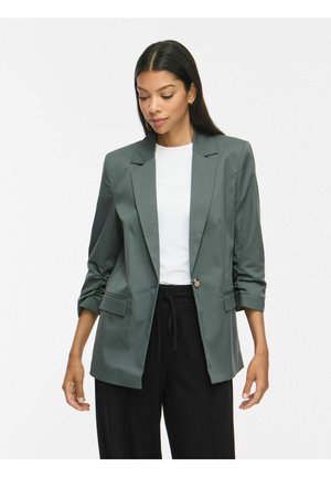 Femme portant un blazer vert foncé sur une chemise blanche et un pantalon noir, avec les manches du blazer remontées de façon décontractée, regardant légèrement vers le bas.