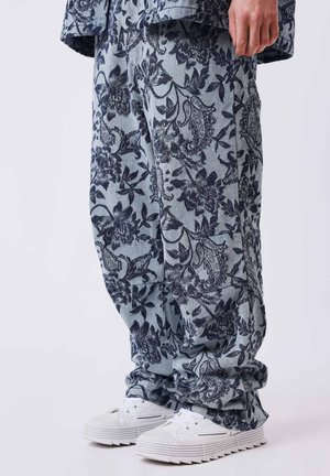 Pantalones holgados con estampados florales oscuros y de paisley sobre un fondo gris claro, combinados con zapatillas blancas con suelas gruesas.