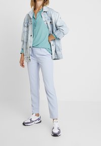 Pantalon bleu clair ajusté, blouse turquoise, et veste en jean délavée associée à des baskets blanches et violettes. Design simple avec des motifs minimalistes.