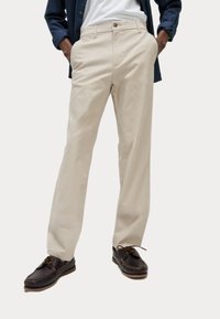 Beige Chinos aus Baumwolle, mit geradem Schnitt, vorderen Taschen und einem einzelnen Knopfverschluss, kombiniert mit dunkelbraunen Bootsschuhen.