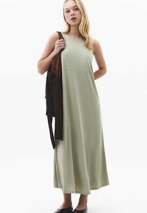 Giovane donna con capelli biondi lisci che indossa un abito lungo verde chiaro senza maniche, tiene una giacca marrone su una spalla, scarpe nere.