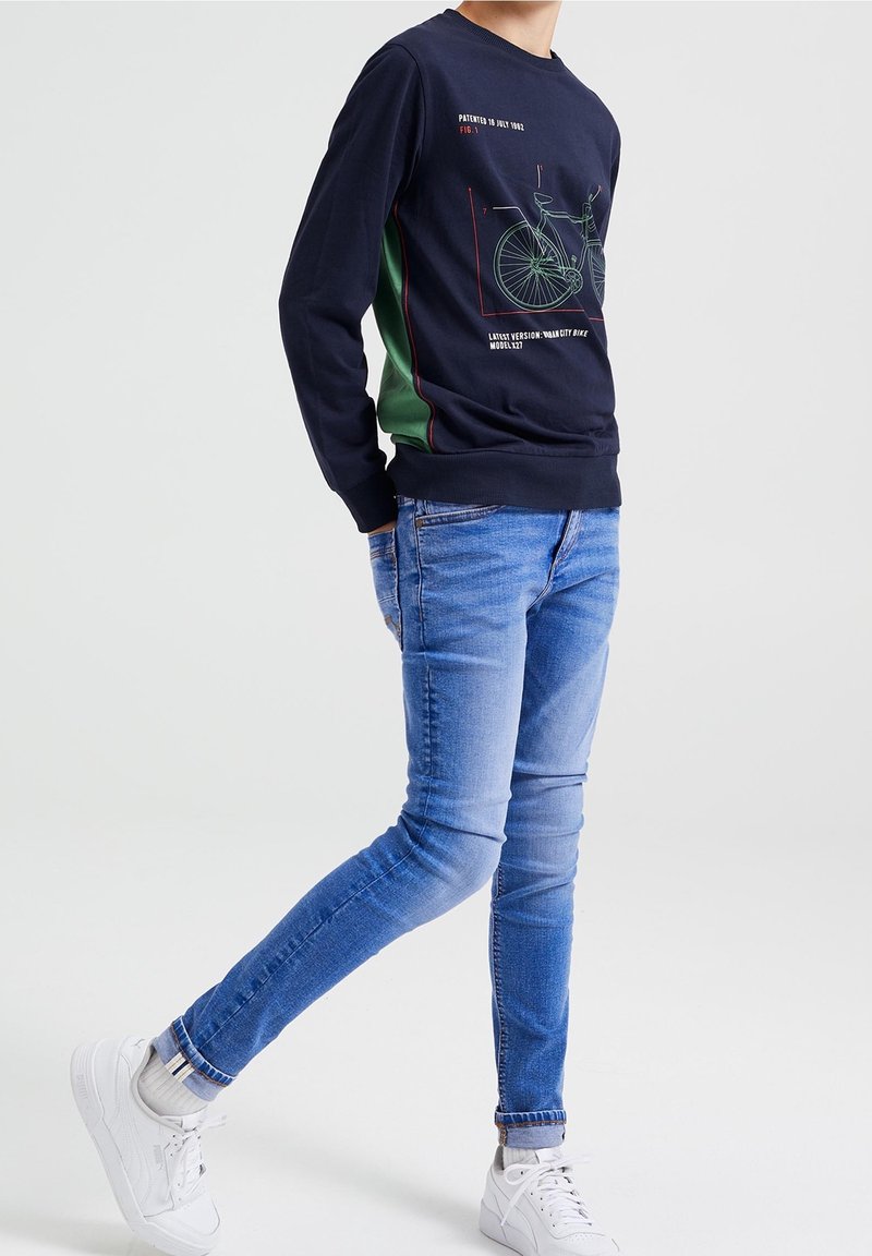 Sudadera azul marino con gráfico de bicicleta y texto, paneles laterales verdes, combinada con jeans de corte ajustado azul claro y zapatillas blancas.