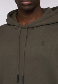 Olivgrön hoodie med en texturerad tyg, snoddar med svarta tippar och en liten broderad logotyp på bröstet.