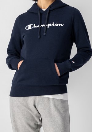 Person iført en marineblå Champion hættetrøje med forlommer og hvid Champion-logo, kombineret med lysegrå træningsbukser.