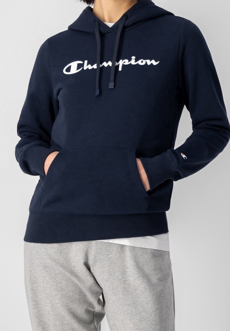 Personne portant un sweat à capuche Champion bleu marine avec une poche avant et un logo Champion blanc, associé à un pantalon de survêtement gris clair.